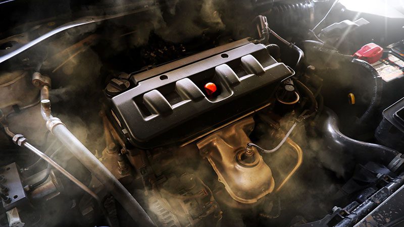 Close-up of a damaged car engine with visible smoke, suggesting overheating or internal engine failure