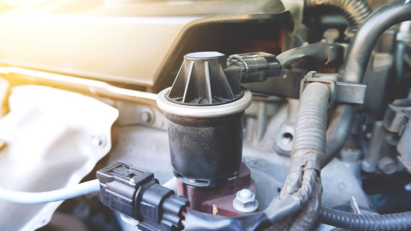 Close-up of an old EGR (Exhaust Gas Recirculation) valve in a car engine compartment, showing its role in reducing carbon monoxide emissions.
