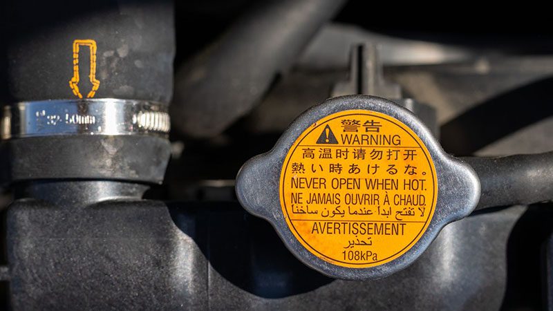 Radiator cap with warning labels and attached hose under the open hood of a car.