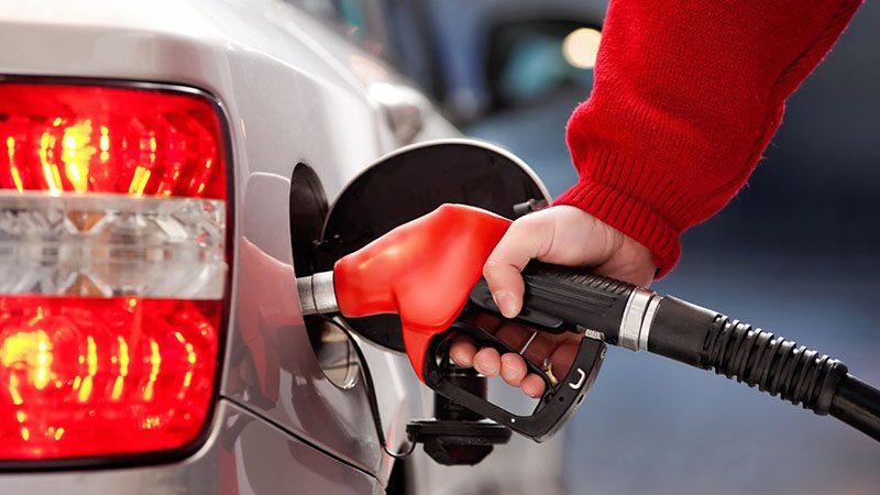 Close-up of a gas station nozzle refueling a large tanker truck, showing fuel hose connection and metal fuel tank.