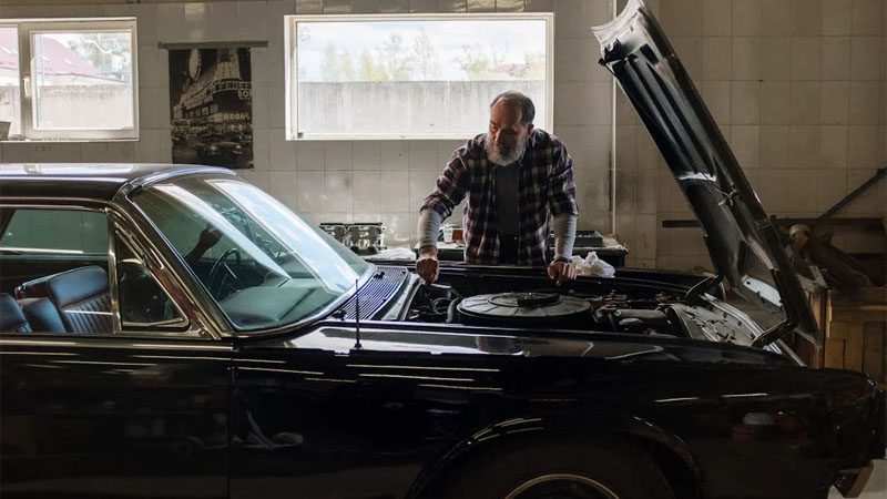 A bearded mechanic examines a classic car engine in a well-lit auto repair shop.