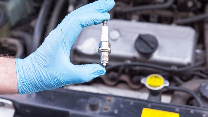 Auto mechanic wearing protective work gloves holds a new spark plug over a car engine during maintenance.
