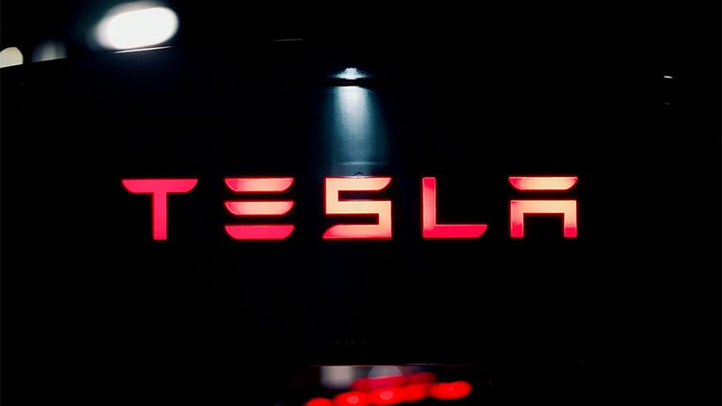 Tesla brand name illuminated at an auto show, featuring dramatic lighting effects and event atmosphere.