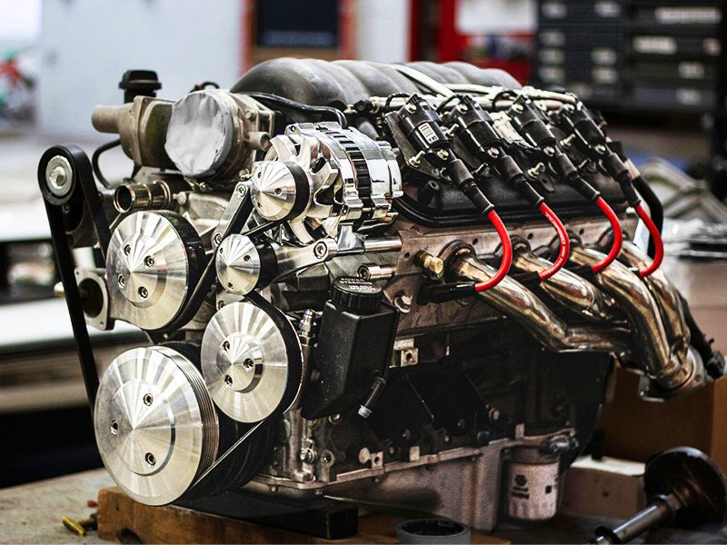 Close-up of a car engine with a vehicle in the background inside a garage, highlighting engine size and setup.