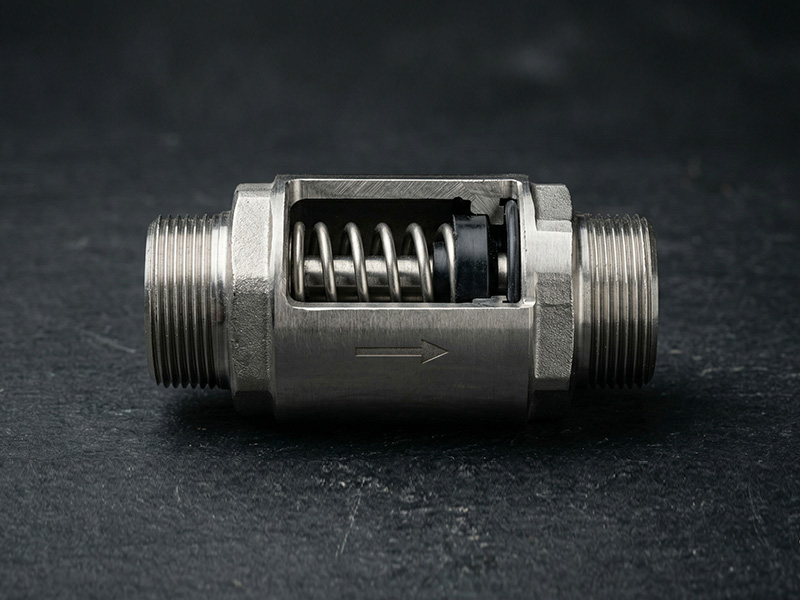 Inline stainless steel check valve with cutaway showing spring-loaded internal mechanism on dark background.