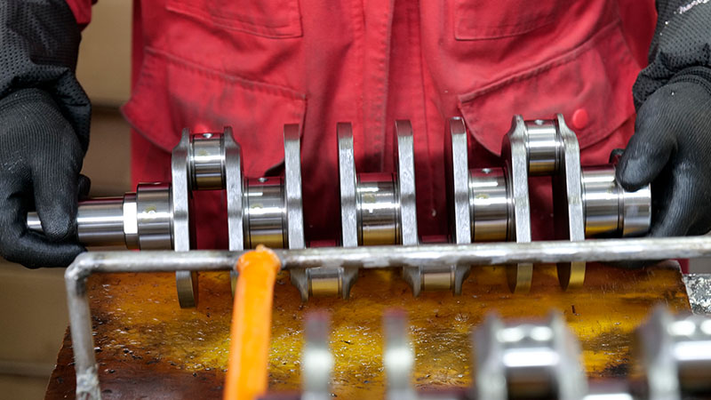 Mechanic holding an automotive engine crankshaft while inspecting its condition and machined surfaces in a workshop.