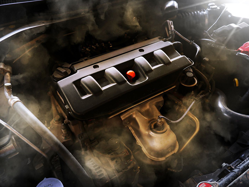 Close-up of a car engine with smoke rising due to blow-by gases, showing signs of engine wear and performance issues.