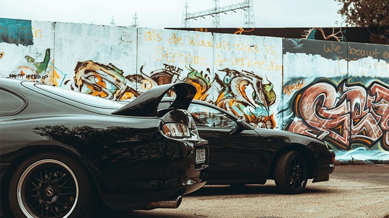 Two retro cars parked against a graffiti-covered wall in an urban setting.